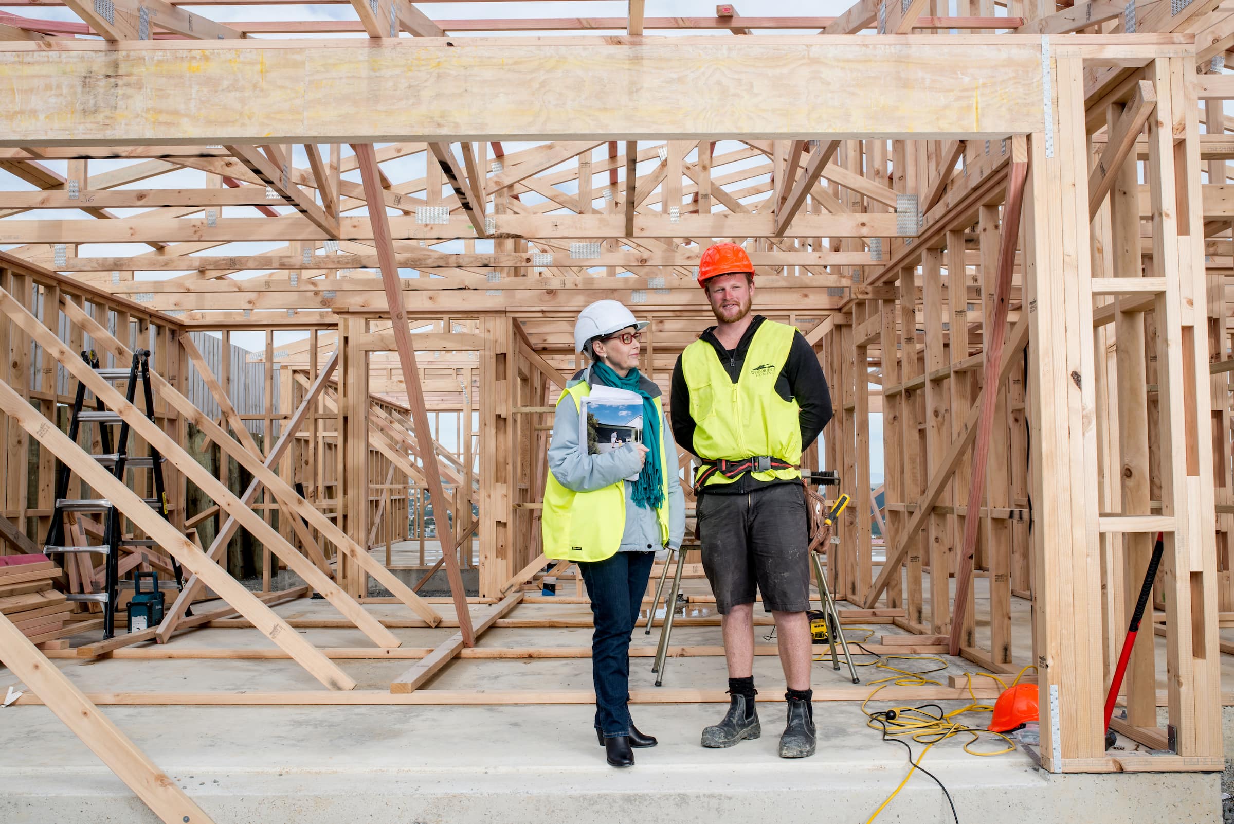 Two architects wearing safety gear stand and talk at a wooden house construction site framed with timber.